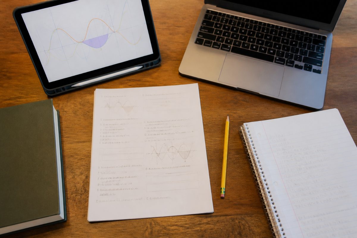 A worksheet, tablet graph, laptop, and notebook are arranged for checking a math problem.