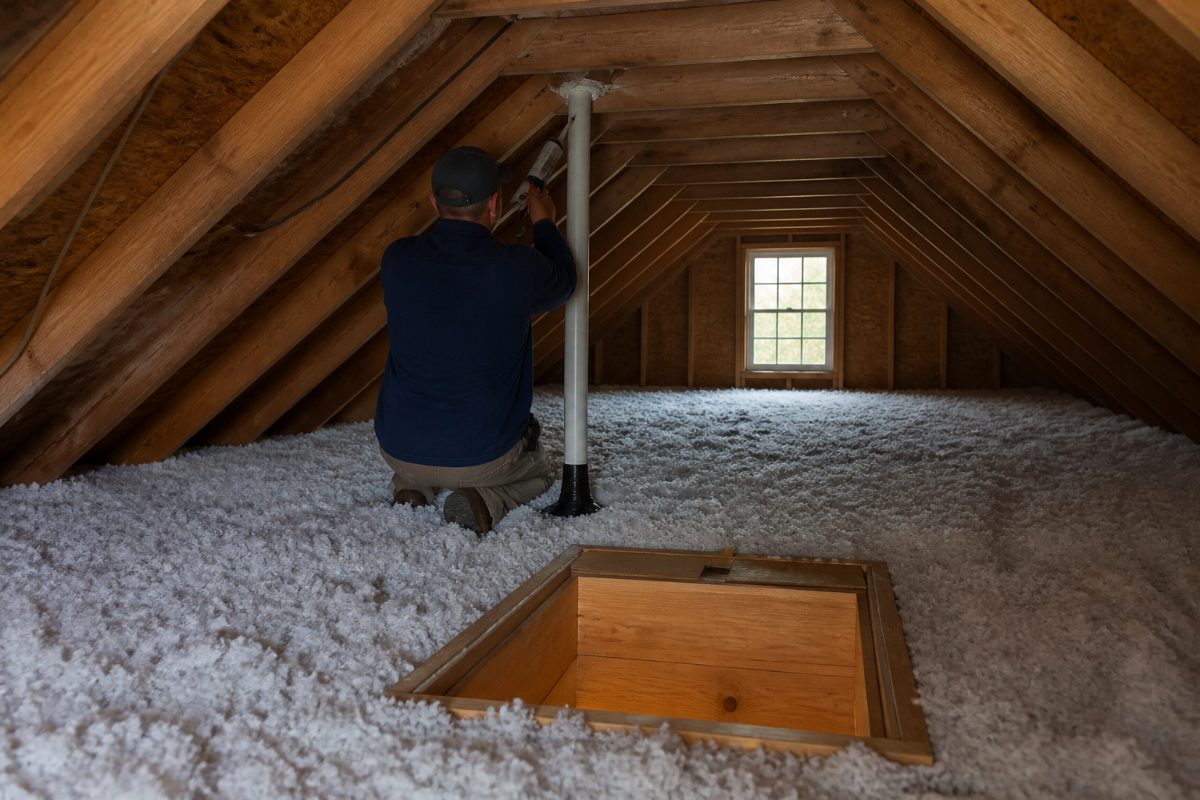 A worker inspects an attic ceiling penetration above the insulation while addressing an air leak.