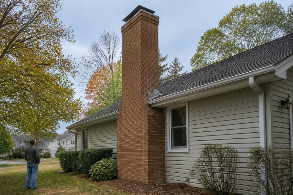 A homeowner views the exterior brick chimney and roofline from the ground during a fall inspection.