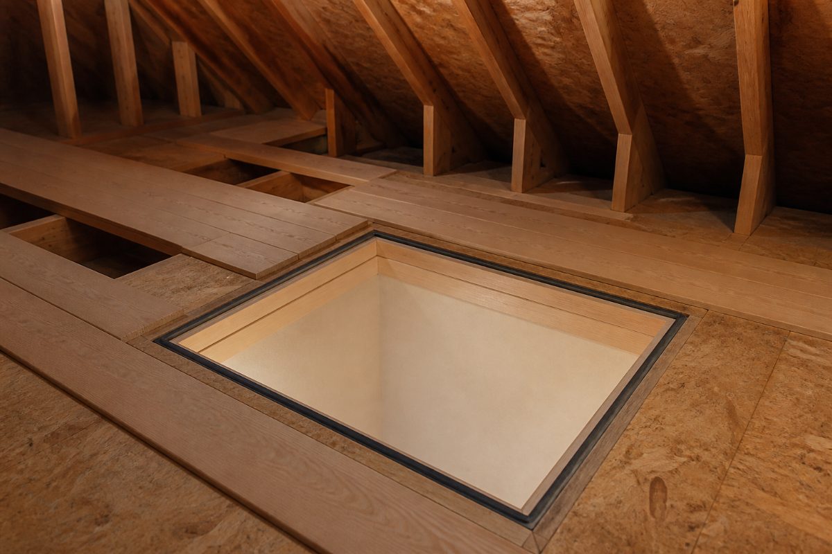 An attic hatch frame and weatherstripping viewed from an attic walkway made of boards across joists.