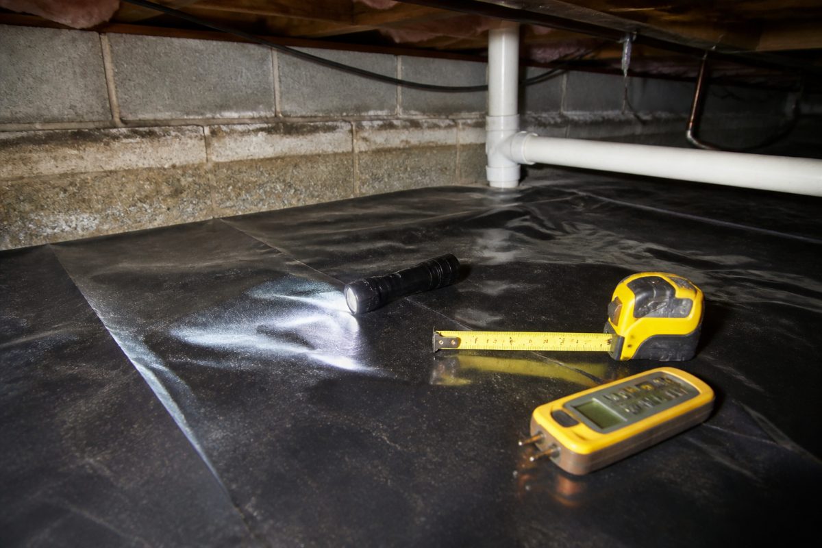 Inspection tools rest on a crawl space liner near a foundation wall and utility pipes.