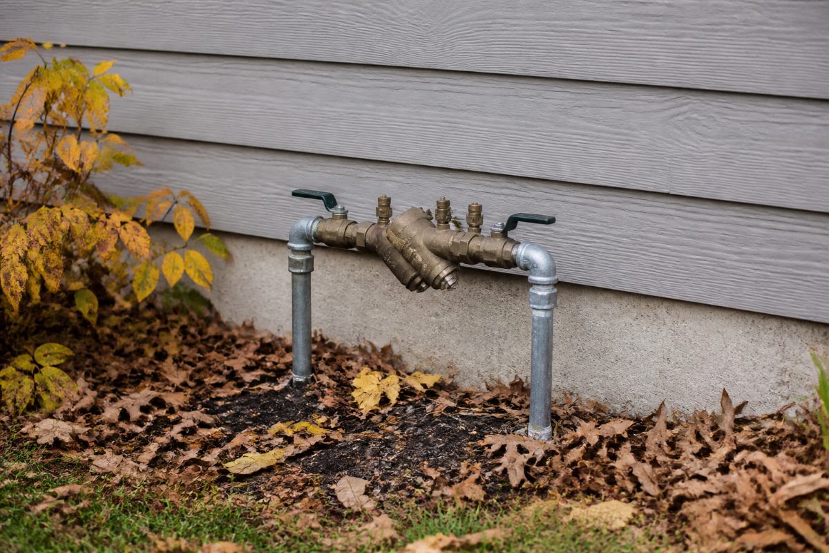 An outdoor irrigation backflow assembly beside a home foundation before winter.