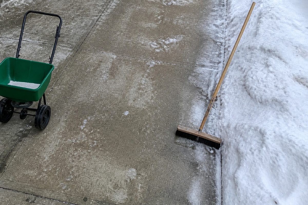A small spreader and broom sit beside a narrow icy strip on a cleared driveway.